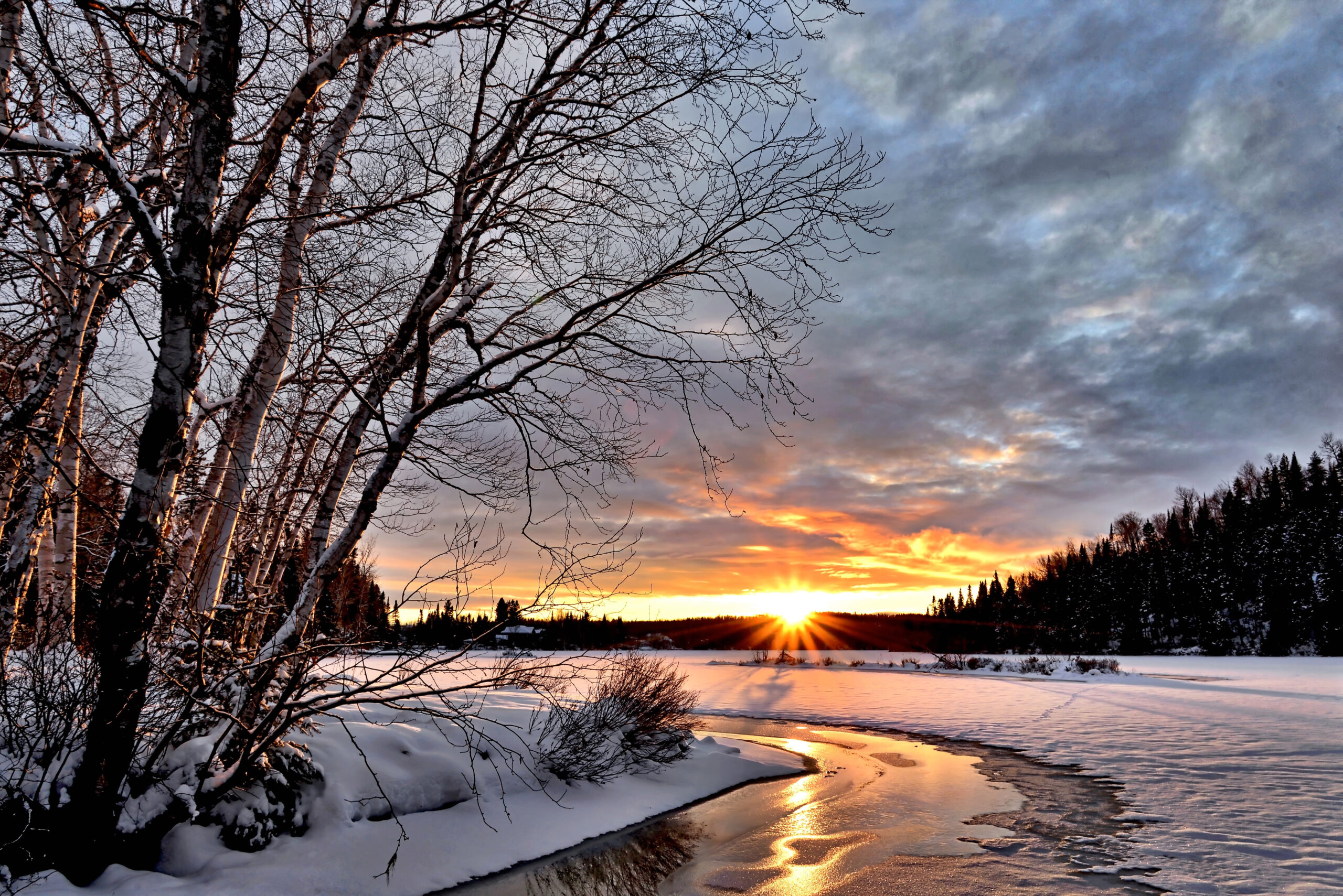 sun setting over a snowy winter landscape with trees and a creek
