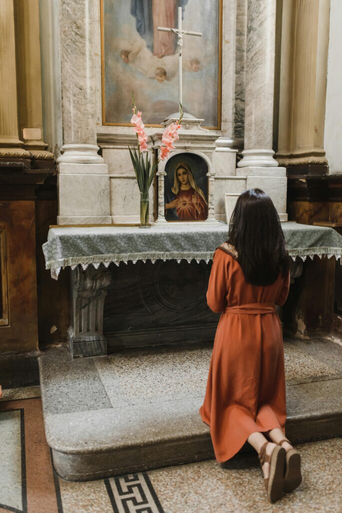 A young woman praying in front of a private altar with an image of the Immaculate Heart of Mary and a vase of flowers on it.