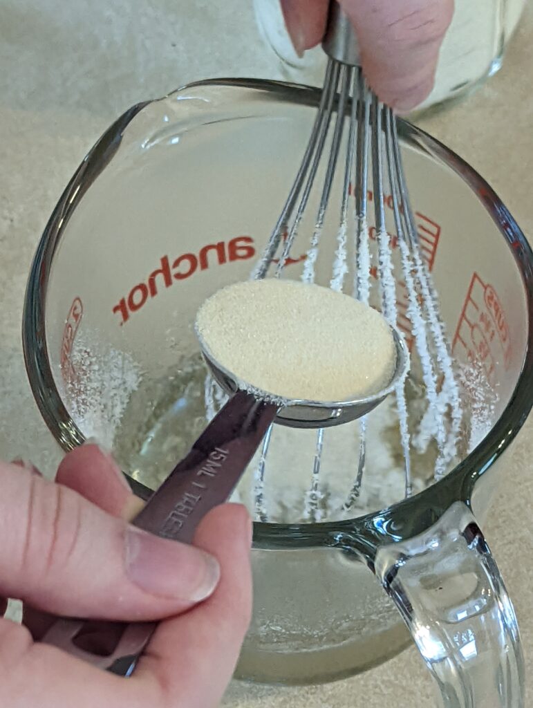 a tablespoon of gelatin being poured into a measuring cup with hot water and a whisk in it